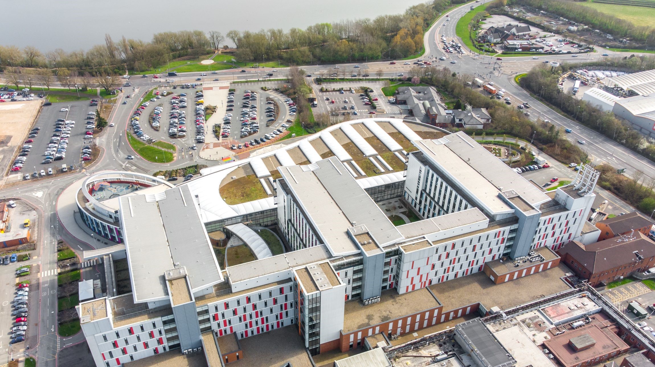 Aerial view of a multistorey hospital building and its car park which are located next to a lake.